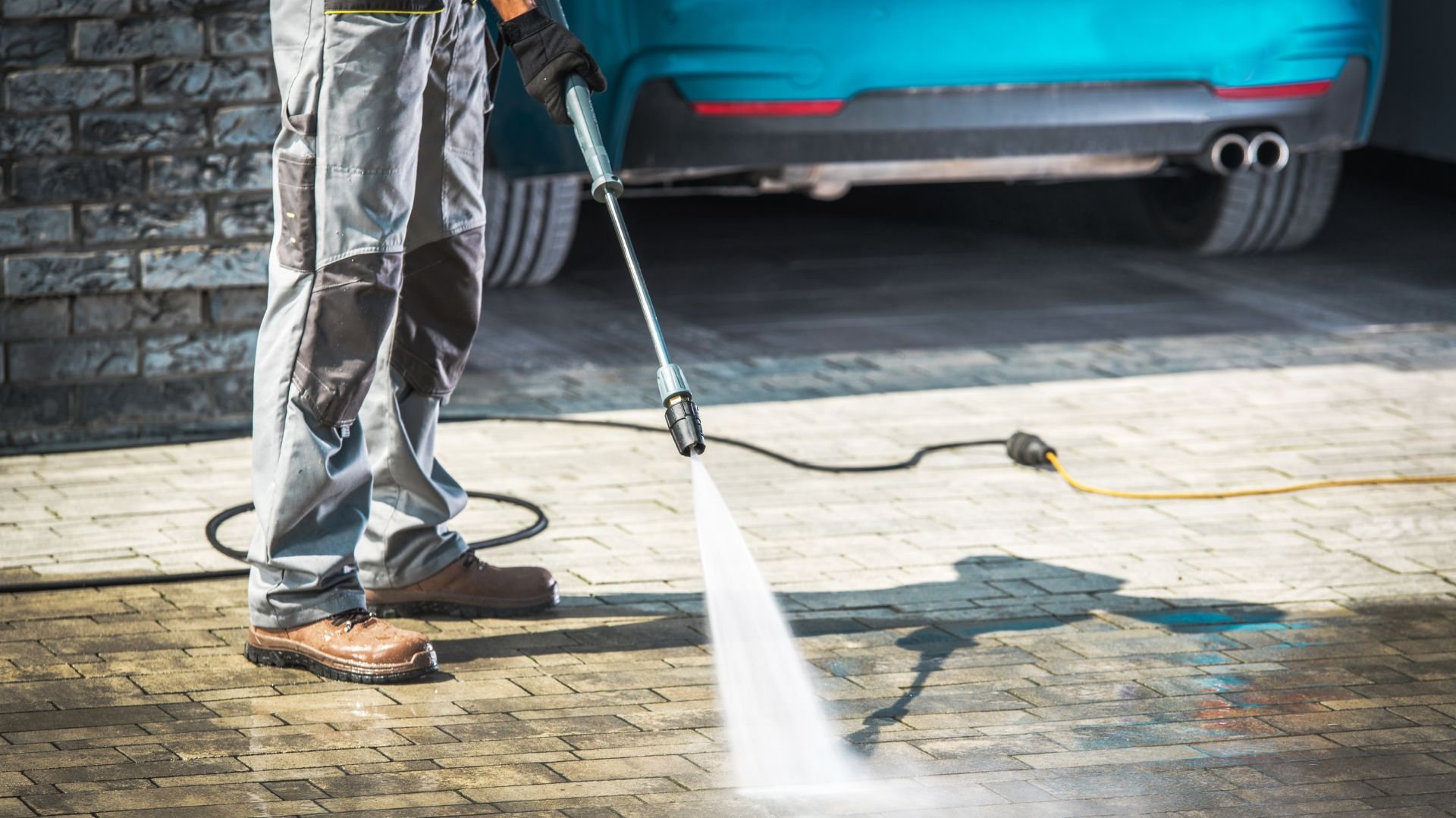 Pressure washer cleaning brick pavement near parked blue car